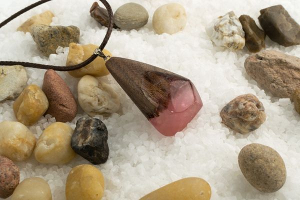 Speckstein trifft Kreativität – Schmuck aus Naturmaterialien Handgemachter Specksteinanhänger mit rosafarbenem Harz und Holz, präsentiert auf weißem Kies mit bunten Natursteinen.